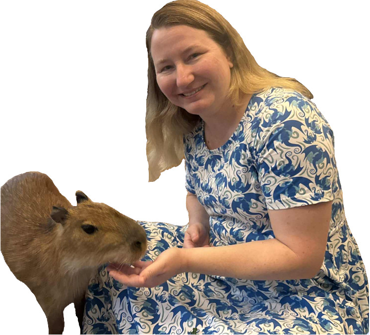 A blond girl in a blue and white dress sitting and feeding a capybara